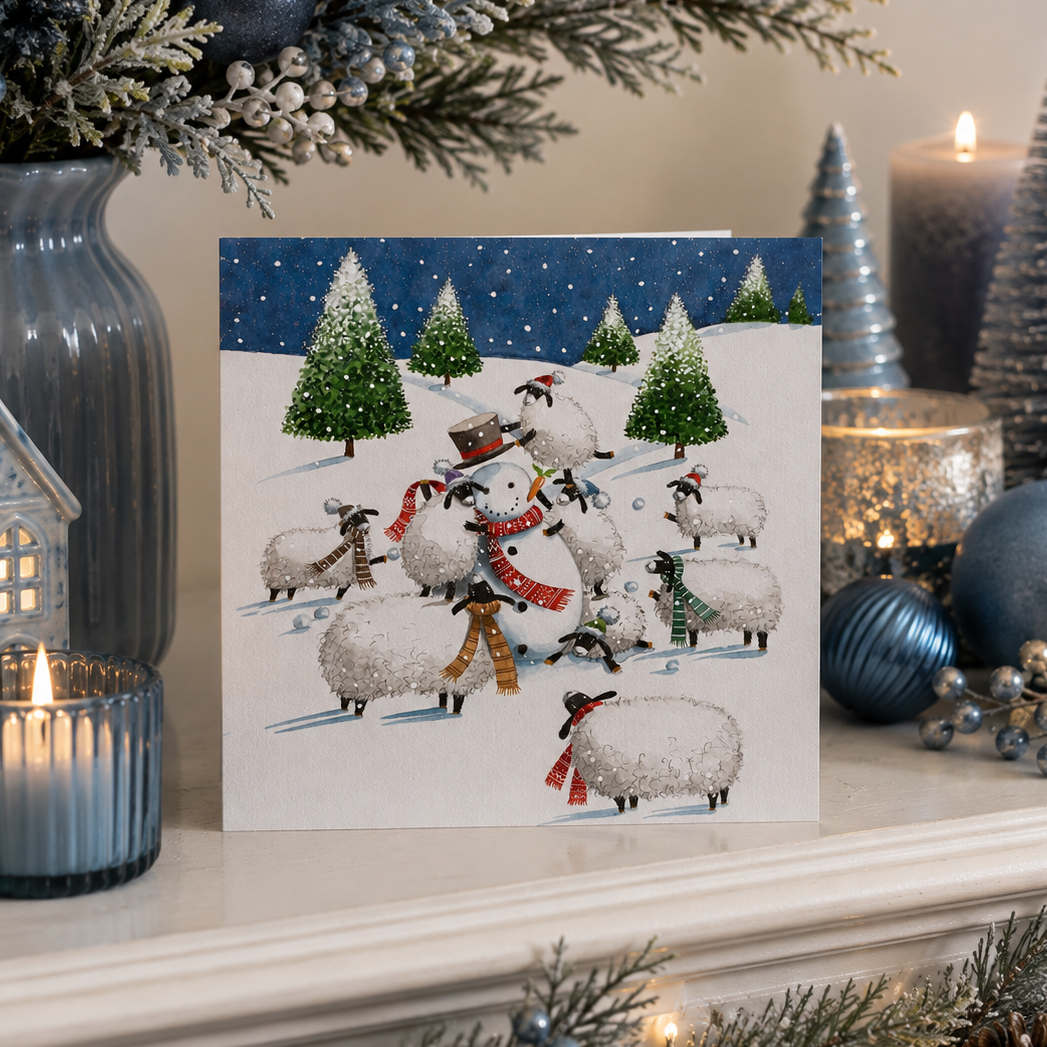 A square greeting card featuring a winter scene with sheep, snowmen, and evergreen trees, displayed on a decorated Christmas mantel with candles, ornaments, and greenery.